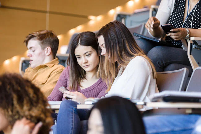 undergrads in a lecture hall, two are leaning in to each other to look at a cellphone screen